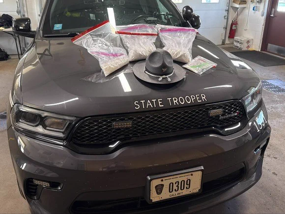 A State Trooper cruiser with several bags of cocaine and a trooper hat displayed on its hood, inside a garage area.