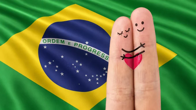 Dois dedos com rostinhos sorridentes desenhados aparecem abraçados sobre um pequeno coração vermelho, com a bandeira do Brasil ao fundo.