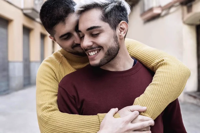 Dois homens sorriem carinhosamente ao ar livre, enquanto um abraça o outro por trás de forma afetuosa.