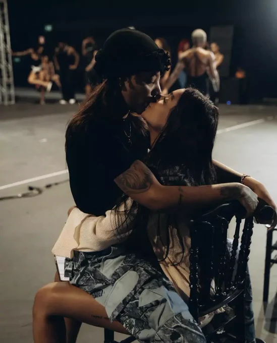 Um casal lésbico se beija em um espaço escuro de bastidores ou apresentação, com outras pessoas desfocadas ao fundo.