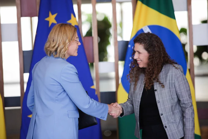 EVP Henna Virkkunen and Minister Esther Dweck shake hands and smile following the signing ceremony. They are standing in front of Brazilian and European Union flags.