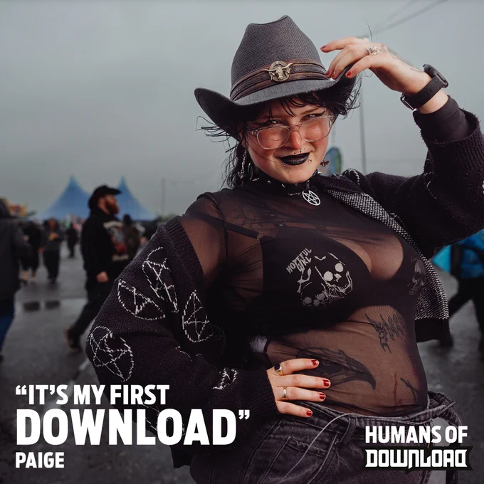 Person wearing a sheer black top with skull designs and a gray hat, posing with their hand on their hat at Download Festival.
