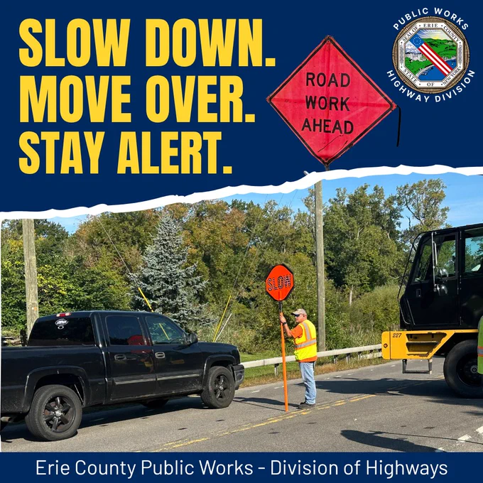 Work zone safety featuring the message “Slow Down. Move Over. Stay Alert.” A highway worker holds a stop/slow paddle while directing traffic near road construction equipment. The Erie County Public Works Division of Highways logo appears in the upper right corner.