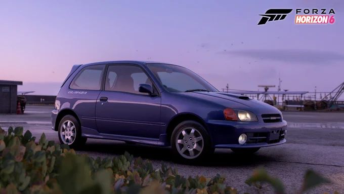 Un coche compacto Toyota Glanza azul está estacionado sobre una superficie pavimentada en una zona abierta con poca vegetación en primer plano. El cielo está despejado, lo que sugiere una hora temprana de la mañana o al final de la tarde. El logotipo de "Forza Horizon 6" es visible en la esquina superior derecha.