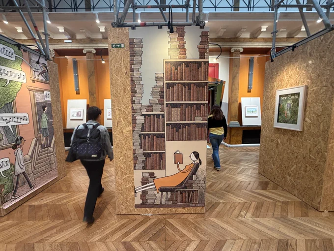 A photograph of a library containing large and small prints of cartoons by Tom Gauld.