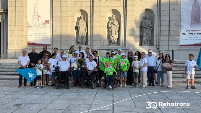 Grupo de personas, incluidas varias en sillas de ruedas, posando frente a un monumento con estatuas, con banderas y flores.