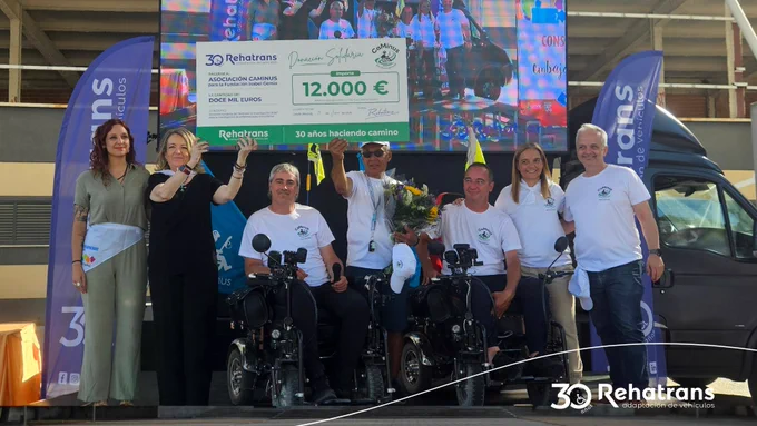 Personas posando con un cheque solidario de 12.000 €; tres usuarios en scooters de movilidad y pancartas de Rehatrans.