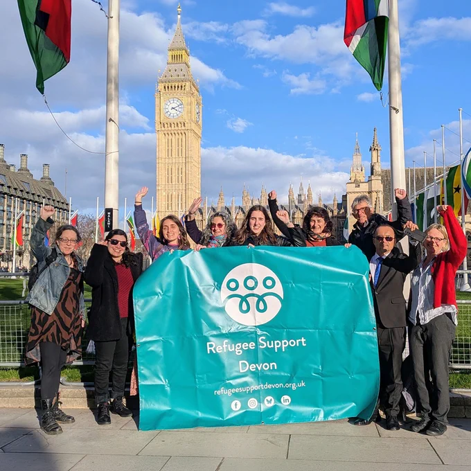 A group of nine people stand outdoors holding a large turquoise “Refugee Support Devon” banner, raising their fists and hands in celebration. Behind them are the Houses of Parliament and Big Ben in London, with flags lining the square under a partly cloudy sky.