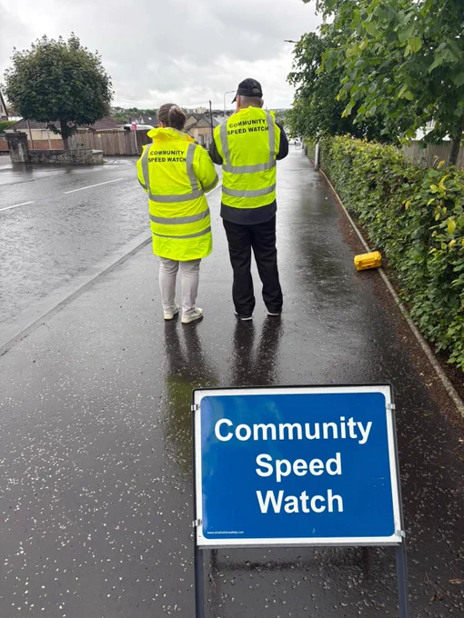 Two people in 'Community Speed Watch' jackets stand on a wet pavement next to a road, with a sign in the foreground.