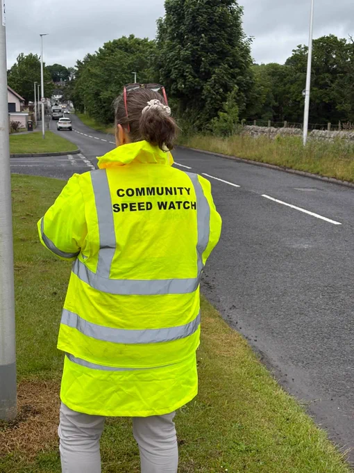 Person in a yellow "COMMUNITY SPEED WATCH" jacket standing by a road.
