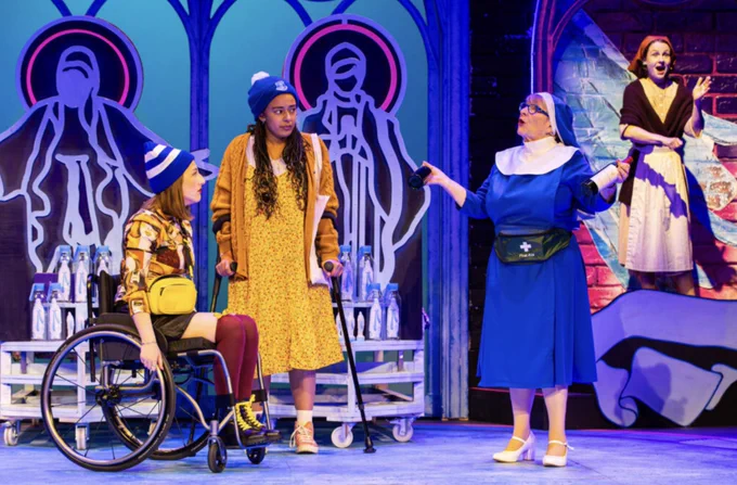 Two people , in bright clothes and both with white and blue bobble hats and one a wheelchair user and one with a stick, are listening to a woman dressed as a Nun talk, her arms outstretched. Behind them a BSL interpreter. They are on a stage. Rose (Maisy Gordon), Helen (Sophia Adzoa-Moore) and Sister Mary (Pauline Daniels). 