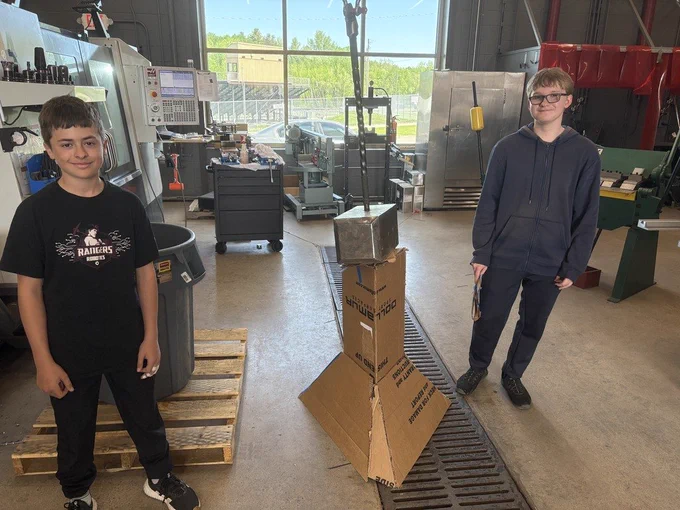 two male students standing next to their carboard design that can hold a construction hammer