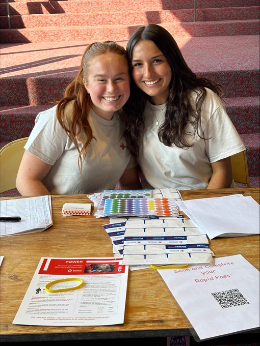 Two individuals sitting at a table with various materials, including a color chart, pamphlets, and a QR code sign. The table appears to be set up for an event or activity, possibly informational or promotional.