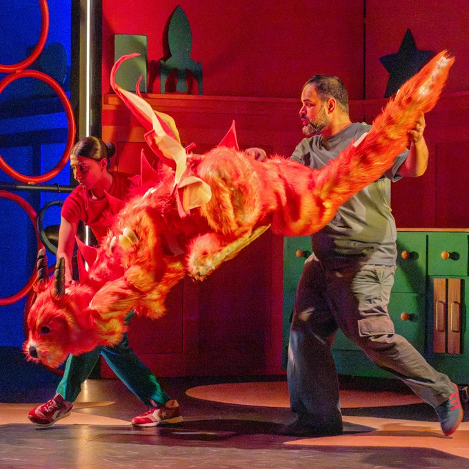 Performers on stage manipulating a large orange and pink dragon puppet. A colourful set with circular props and a cupboard is in the background.