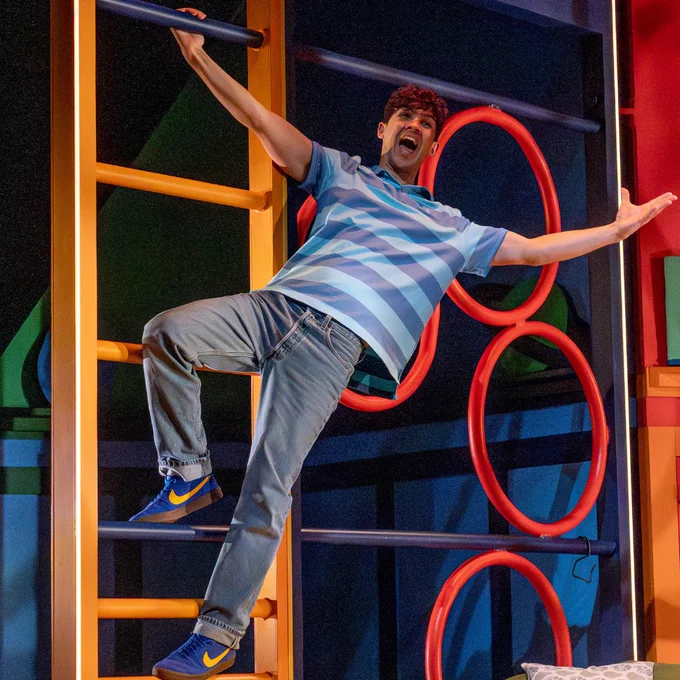 A person playfully climbs on a colourful structure with circular and ladder elements. They are wearing a blue striped shirt, jeans, and blue sneakers with Nike logos. The background features vibrant colours and geometric shapes.