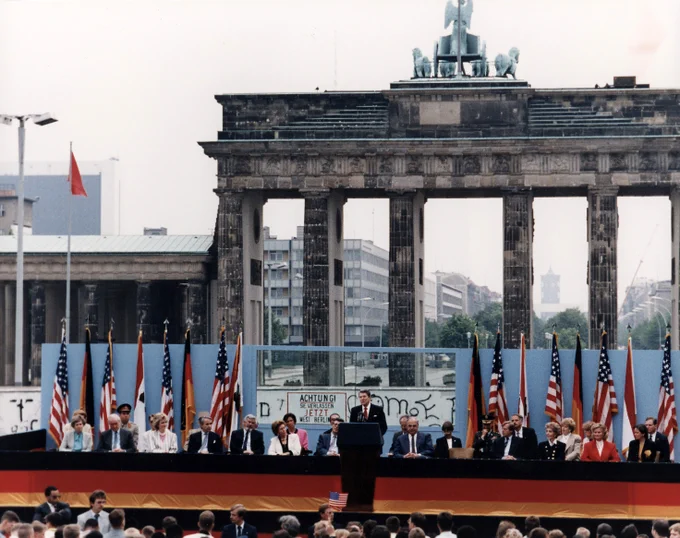A formal event with officials seated before the Berlin Wall and Brandenburg Gate, decorated with American flags and a large crowd in front. President Reagan stands in the center of the image at a podium.