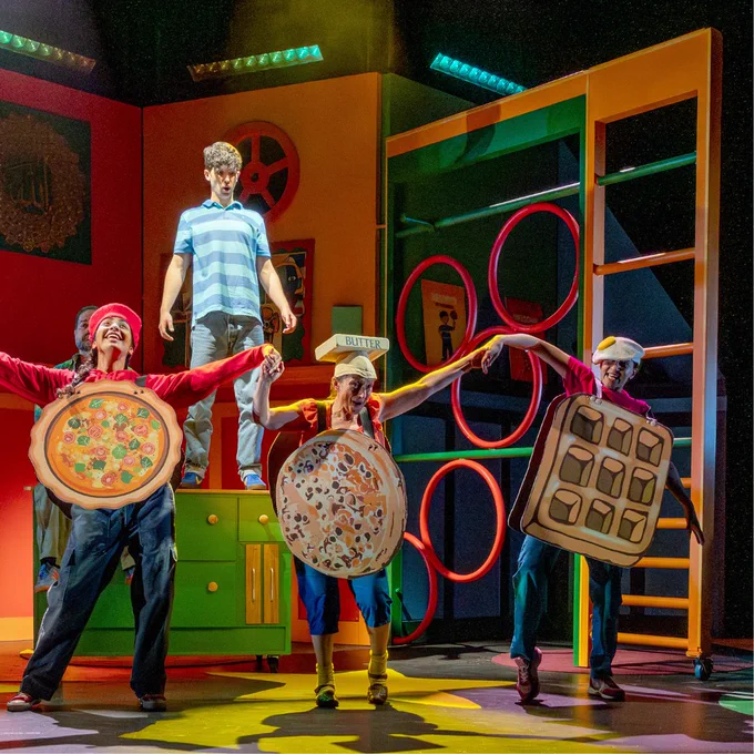 A theatrical performance featuring a group of performers on stage. Two performers are dressed as pizzas, and another as a waffle. A fourth performer stands elevated in the background wearing a striped shirt. Brightly coloured set pieces and props surround them, adding to the lively scene.