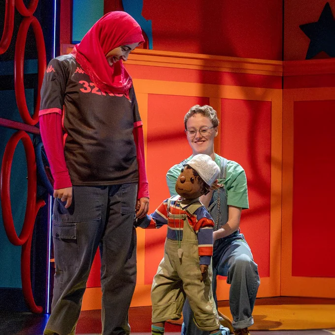 A person wearing a colorful outfit and headscarf looks down and smiles at a puppet of a child holding their hand. Another individual, seated nearby, interacts with the puppet, which is dressed in a striped shirt and overalls. The background features vibrant red and orange panels with large stars.