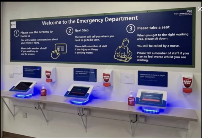 Screen shot The Times 11/06/2026 photo of Kings College Hospital  ED  with 3 tablets on stands with uv light under each on a white shelf Alcohol hand gel on wall above each Large Blue sign white text with instructions on what to do on cream wall