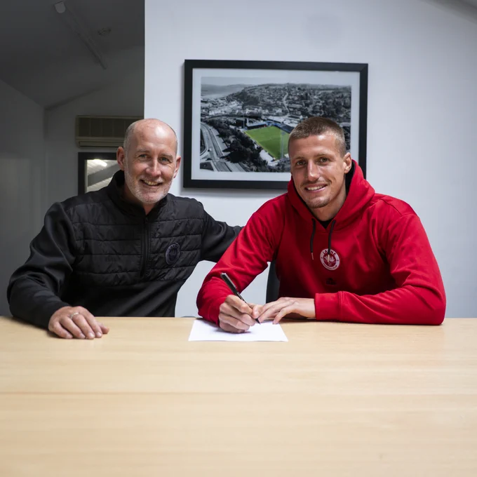 Two men are sitting at a table, one signing a document. The setting has a professional and upbeat mood.