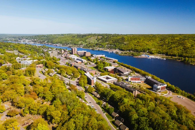 Aerial view of a riverside town with dense green trees, buildings, and a winding road under a clear blue sky.