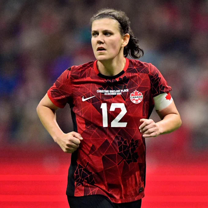 Christine Sinclair runs on a soccer field wearing Canada’s red number 12 jersey, black shorts, and a white captain’s armband with a red maple leaf. The stadium crowd is blurred in the background, with a bright red field-level barrier across the lower part of the image.

