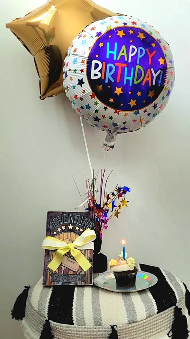 Happy Birthday balloons above a chocolate cupcake with a lit candle, and a copy of Adventura, wrapped with a green and white bow.