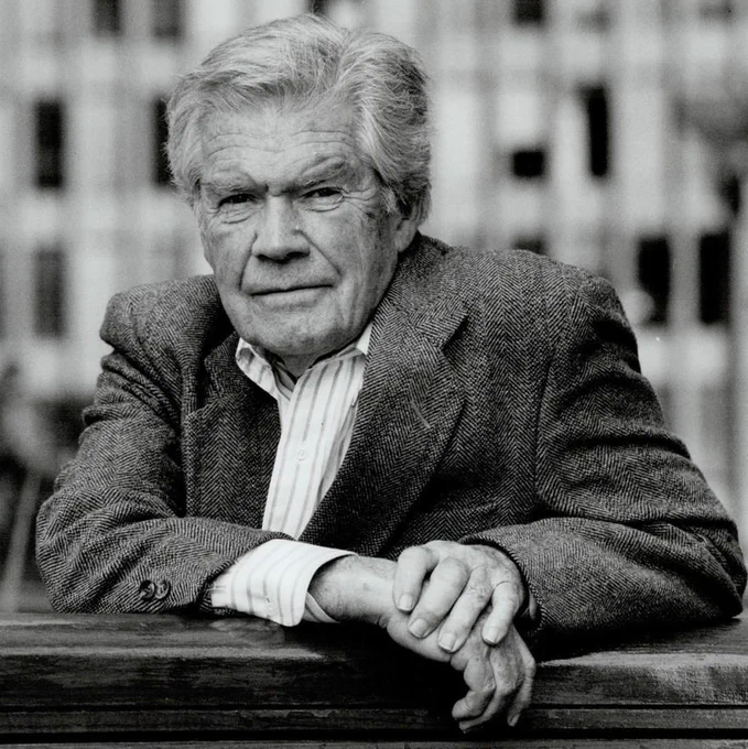 Scott Young leans on a wooden railing in a black-and-white portrait, wearing a textured blazer over a striped shirt. His hands are folded in front of him, and he looks directly at the camera. The background is softly blurred with vertical shapes from nearby buildings or structures.
