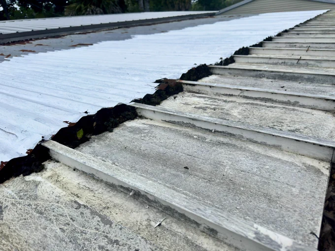 Wide view of a hurricane-damaged metal roof on a mobile home in Florida. New white roofing panels have been installed on the upper section, while older lower panels still show rust and weather damage. View of the transition between newly installed white metal roofing and older rust-stained roof panels. The damaged section remains exposed and in need of repair before storm season. Close-up of the mobile home’s roof framing beneath a lifted metal panel. A weathered support beam and exposed fastener highlight the unfinished repairs and vulnerability to leaks and hurricane winds. Panoramic view across a partially repaired mobile home roof in Florida. Bright white metal panels contrast with rusted sections that still require replacement. Large trees and summer clouds fill the background.