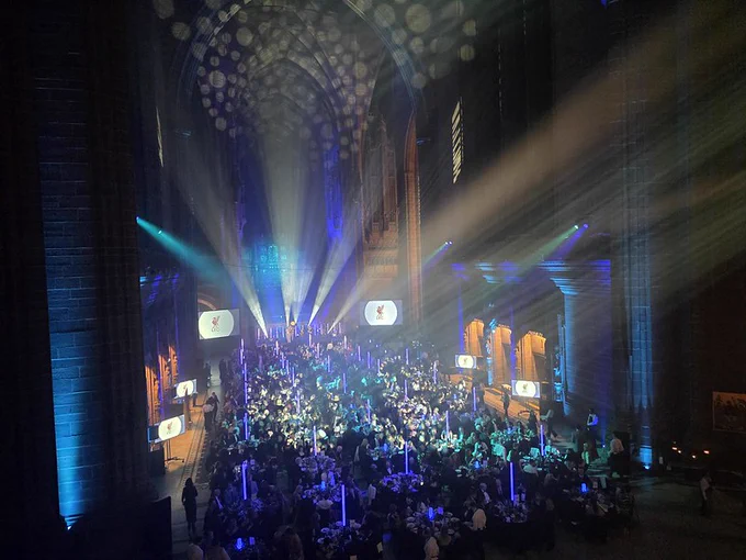 A large, cathedral-like hall set up for a formal evening event, with long banquet tables filled with seated guests. Blue and white stage lighting beams downward from a high, arched ceiling, creating dramatic light patterns. Two large screens near the front display a logo, while the crowd fills the space from foreground to background.