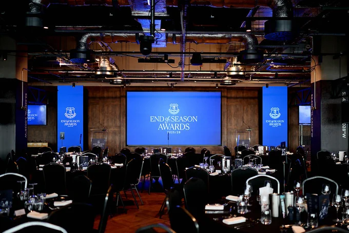 An indoor awards event setup with round dining tables neatly arranged in front of a stage. A large central screen displays the text “END OF SEASON AWARDS 2025/26,” flanked by two vertical banners with the same branding. The room features dark chairs, table settings with glassware, and overhead industrial-style lighting.