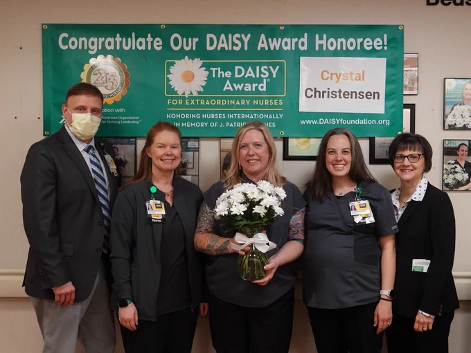 A group of five individuals stands in front of a green banner congratulating a DAISY Award honoree for extraordinary nurses. The honoree, holding a bouquet of white flowers, is flanked by colleagues. The banner prominently displays the name "Crystal Christensen." The group appears joyful, and they are wearing identification badges, with one individual also wearing a face mask.