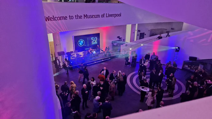 A modern museum atrium hosting a networking event, viewed from an upper level. Groups of people stand and talk around tall tables. At the far end, a small stage with a screen reads “50 YEARS” alongside a logo, while colorful lighting casts purple and blue tones across the open space. The wall above displays the text “Welcome to the Museum of Liverpool.”