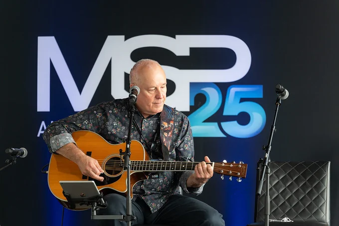 Performer seated on a small stage, playing an acoustic guitar into a microphone. A large backdrop behind the stage displays the text “MSP 25,” with additional microphones and equipment visible around the performer.