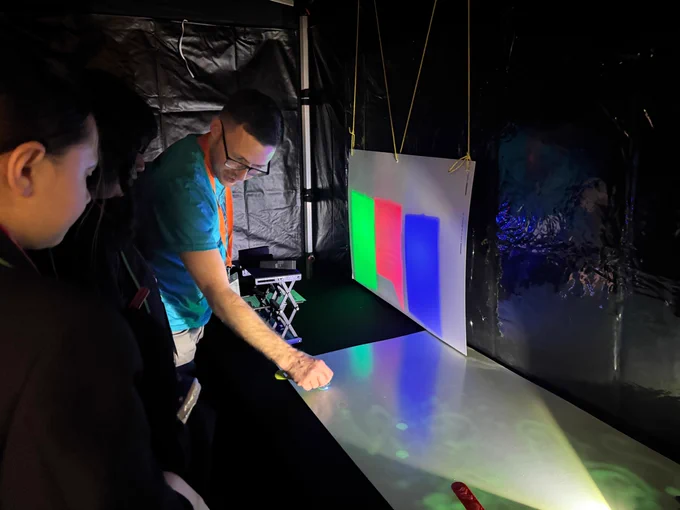 A person wearing glasses and a teal shirt is demonstrating colored lights at the National Science and Media Museum. A projection with red, green, and blue lights is shown on a white surface, with several people observing the setup.