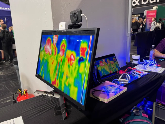 A colorful thermal image displayed on a monitor at an event booth. The setup includes a laptop, some papers, an infrared camera, and equipment cases. People in the background are interacting with other booths.