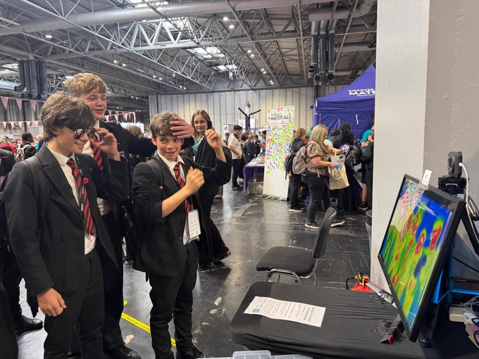 A group of students in school uniforms are enjoying an interactive exhibit. They are looking at a screen displaying colourful, abstract visuals. The event is busy, with people and various stands visible in the background.