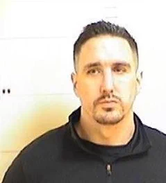 Mugshot of a man with short dark hair and facial hair wearing a black jacket.