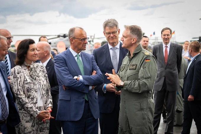 Auf dem Bild sieht man Bundesverkehrsminister Patrick Schnieder zusammen mit Bundeskanzler Friedrich Merz und Bundesforschungsministerin Dorothee Bär im Gespräch mit einem Mitarbeiter der Luftwaffe auf der ILA 2026 in Berlin.