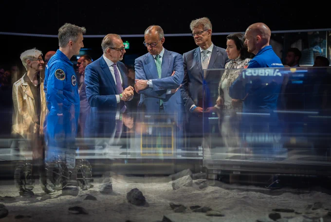 f dem Bild sieht man Bundesverkehrsminister Patrick Schnieder zusammen mit Bundeskanzler Friedrich Merz, Bundesforschungsministerin Dorothee Bär und Mitarbeitern der European Space Agency, die sich auf der ILA 2026 in Berlin über technologische Entwicklungen in der Raumfahrt informieren. Vor ihnen ist eine Weltraumlandschaft nachgestellt.