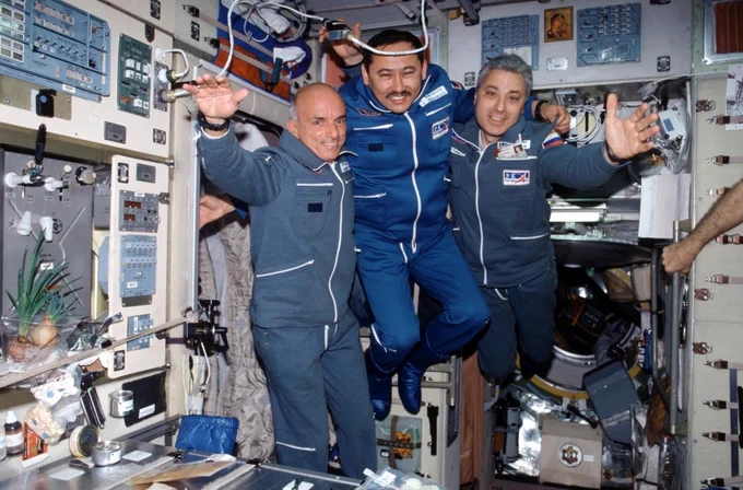  Yuri Baturin (pictured at right next to Dennis Tito and Talgat Musabayev) floats aboard the International Space Station. All three men are wearing flight suits in various shades of blue.