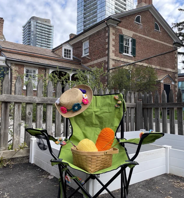 Green folding chair with a basket of colorful yarn and a decorated straw hat, set outdoors near a wooden fence and brick building.