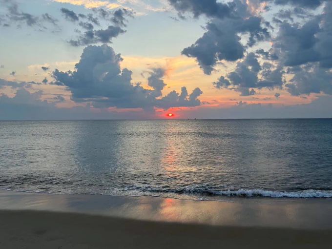A serene beach scene at sunset with a vibrant orange and red sun rise over the horizon. The sky is filled with dramatic clouds reflecting on the calm ocean water, and a small wave gently laps the sandy shore.