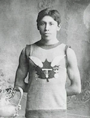 Historic black-and-white portrait of a young Indigenous athlete standing and posing with a trophy. He wears a sleeveless athletic jersey featuring a large maple leaf emblem with wings across the chest, suggesting participation in an early Canadian sporting competition. His hair is neatly parted, and he stands confidently with one hand behind his back while holding the trophy in the other. The photograph appears to date from the early 20th century and reflects both athletic achievement and Indigenous participation in Canadian sport during a period when Indigenous peoples faced significant social and political barriers.

Context: This is a well-known photograph of Tom Longboat (Cogwagee), the celebrated Onondaga long-distance runner from the Six Nations of the Grand River. Widely regarded as one of Canada’s greatest athletes, Longboat won the 1907 Boston Marathon and set numerous professional distance-running records. He became an important Indigenous sports pioneer whose accomplishments