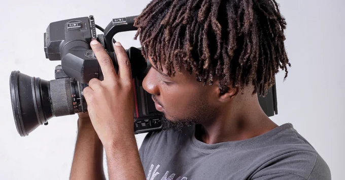 A person holding a professional video camera, looking through the viewfinder. They have short, twisted hair and are wearing a dark T-shirt. The background is plain white.