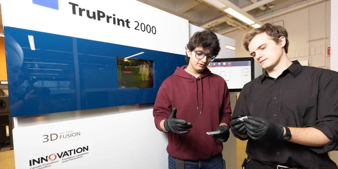 Two students wearing disposable gloves each hold a small   
metal object as they stand in front of a large TRUMPF TruPrint 2000 machine.  