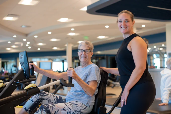 Senior woman using a T6 recumbent cross trainer in a gym while a trainer stands beside her offering support and guidance.