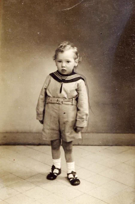 A vintage black and white photograph of a young boy standing in a studio, wearing a sailor-style outfit with shorts and a collared top. The child has curly hair, and is wearing socks with dark shoes.
