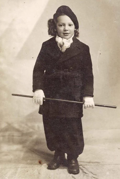 Vintage photograph of a child dressed in a formal coat and scarf, holding a walking stick, standing against a plain backdrop.