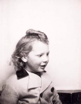 Vintage black and white photograph of a young child in a coat with a buttoned collar and styled hair, looking to the side with a slightly open mouth. The background is plain and light-colored, emphasizing the subject in the foreground.
